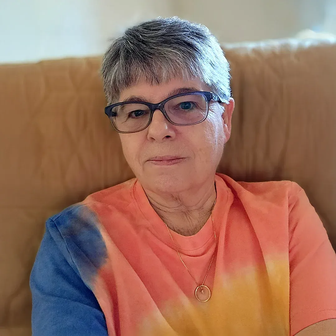 Portrait Of Cindy Baudoin Wearing Glasses And A Colorful Shirt Cindy Baudoin with short gray hair and glasses wearing a colorful shirt while seated indoors